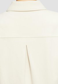 Chemise beige clair avec un tissu texturé, un col classique, et une pince centrale dans le dos pour un détail de design supplémentaire.