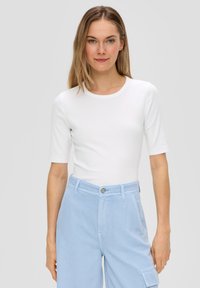 Weißes, eng anliegendes T-Shirt mit kurzen Ärmeln und rundem Halsausschnitt, kombiniert mit hellblauen, hoch taillierten Hosen mit Knopfverschluss und Taschen.