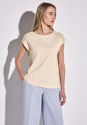 Top corto de color beige claro con cuello redondo y detalles de encaje intrincados en los hombros; combinado con pantalones sueltos de color azul claro.