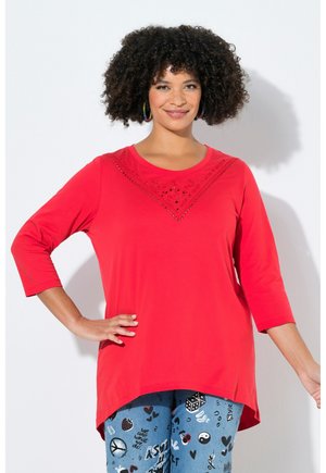 Kobieta o kręconych włosach ubrana w czerwoną bluzkę z długim rękawem z dekoracyjnym dekoltem oraz niebieskie spodnie z różnymi czarno-białymi nadrukami graficznymi.