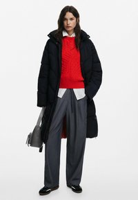 Abrigo tipo puffer en negro, suéter de punto trenzado rojo, camisa blanca de cuello, pantalones de pierna ancha grises y zapatos negros. Los accesorios incluyen un bolso gris.
