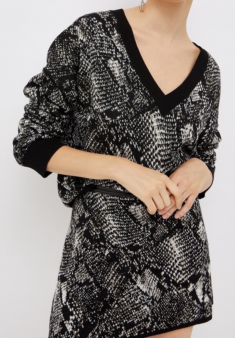 Femme portant une robe-pull à col en V avec un motif noir et blanc, manches longues légèrement froncées aux poignets, et une fine ceinture noire.
