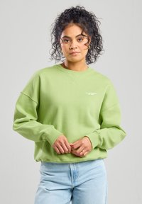 Mint green sweatshirt with a crew neck, relaxed fit, and ribbed cuffs. Features white text logo on the chest. Paired with light blue jeans.
