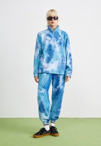 Blå tie-dye fleece genser med glidelås i kragen, kombinert med matchende joggebukser. Myk tekstur, avslappet passform og elastiske mansjetter med hvit-blå aksent.