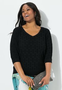 Schwarzes strukturiertes Strickoberteil mit V-Ausschnitt und dreiviertel Ärmeln, kombiniert mit gemusterten Hosen und einer metallischen Clutch.
