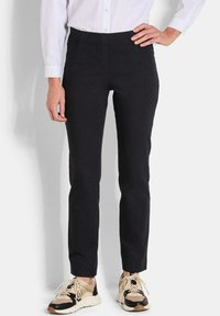 Persoon in zwarte slim-fit broek met beige en zwarte sneakers, staande met één hand op de heup tegen een witte achtergrond.