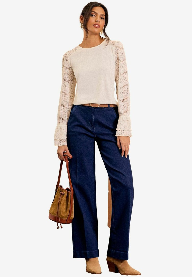 Beige Strickoberteil mit langen Spitzärmel, kombiniert mit dunkelblauen weit geschnittenen Jeans. Brauner Gürtel und hellbraune Stiefeletten, dabei eine braune Bucket-Bag.