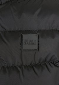 Czarna kurtka puchowa z bliska, z teksturowanej, syntetycznej tkaniny z przeszytym wzorem i kwadratową, wytłoczoną naszywką logo Urban Classics.