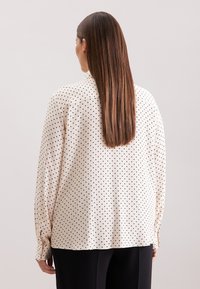 Lange mouwen, crèmekleurige blouse met zwarte stippen. Zacht materiaal, ontspannen pasvorm, knoopdetails op de mouwen en een gladde textuur.