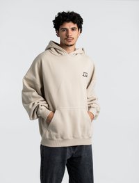 Felpa oversize beige con tasca a marsupio frontale, orlo e polsini a coste, caratterizzata da un piccolo logo sul petto. Indossata con jeans scuri.