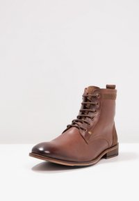 Bottines en cuir marron avec une finition lisse, bout rond, design à lacets et panneau en tissu contrastant sur le côté. Semelle en caoutchouc avec un relief minimal.