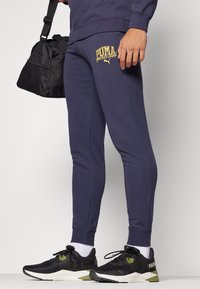 Marineblauwe joggingbroek met een gouden "PUMA sports club" logo, taps toelopend ontwerp, zachte stofstructuur. Gecombineerd met zwarte sportschoenen en een zwarte tas.