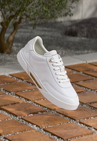 Adidași albi cu logo auriu pe lateral, suspendați deasupra unor plăci de lemn și mici pietrișuri gri, cu o plantă verde neclară în fundal.