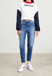 Corte de suéter blanco de punto con mangas azul marino, con el logo "TOMMY JEANS". Jeans de cintura alta azul claro. Zapatillas blancas con detalles rojos.