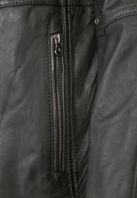 Schwarze Lederjacke mit einer Reißverschlusstasche, glatter Textur und detaillierter Naht entlang der Kanten. Der Reißverschluss hat eine metallische Oberfläche.