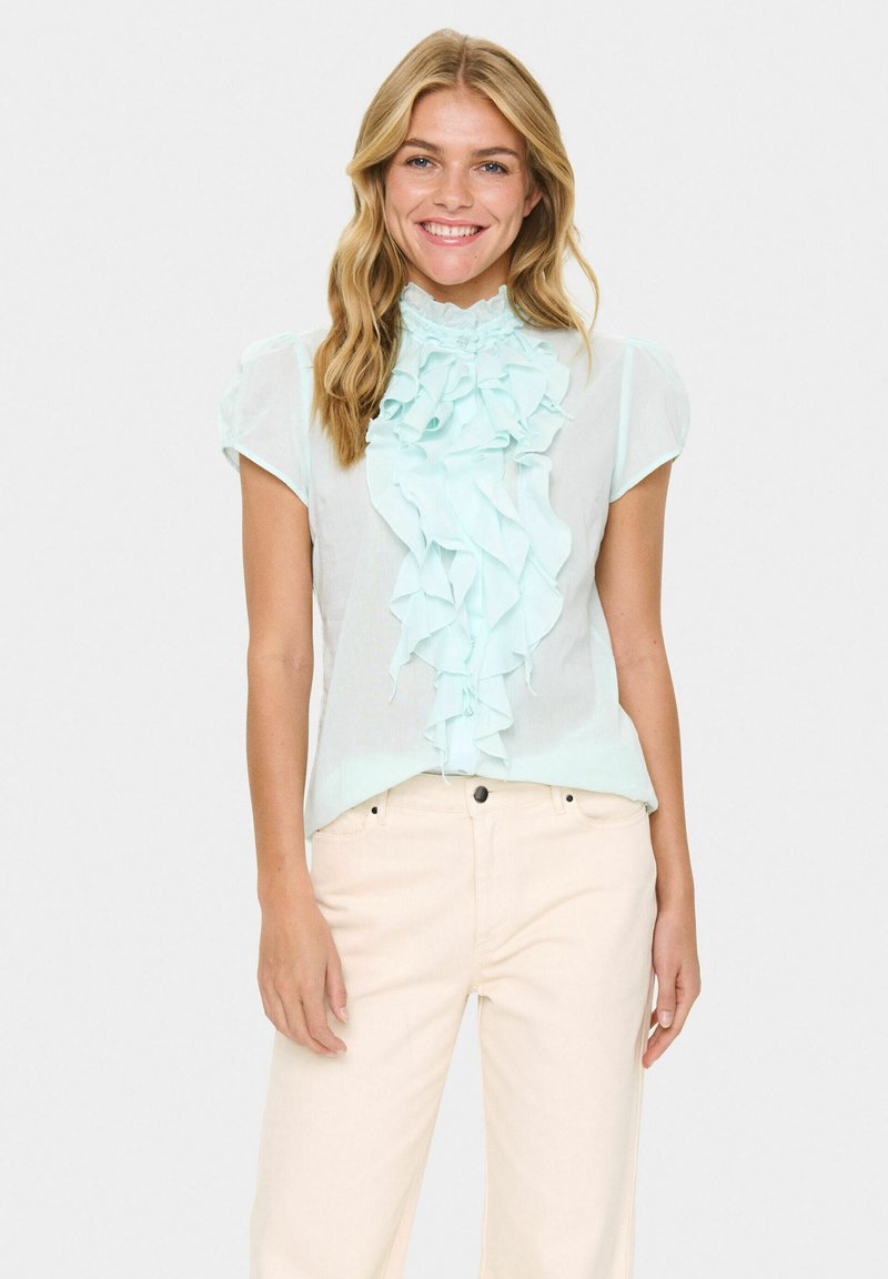 Blusa verde menta chiaro con dettaglio arricciato sul davanti e maniche corte, realizzata in tessuto leggero. Abbinata a pantaloni beige.