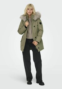 Parka vert olive avec capuche en fausse fourrure, fermeture éclair et bouton, poches latérales, manches longues et coupe décontractée. Pantalons noirs et bottes portés.