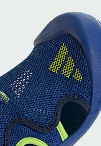 Verkkokudottu urheilujalkine tummansinisenä neonvihreillä yksityiskohdilla. Varustettu teksturoidulla pinnalla, kaarevilla reunoilla ja ilmanvaihtoa varten tarkoitetuilla aukkoilla.