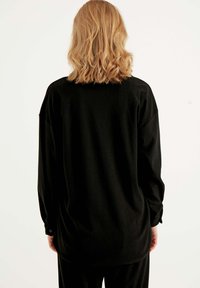 Schwarzes Langarmshirt mit gerippter Textur, das einen lockeren Schnitt und geknöpfte Manschetten aufweist, von hinten betrachtet.