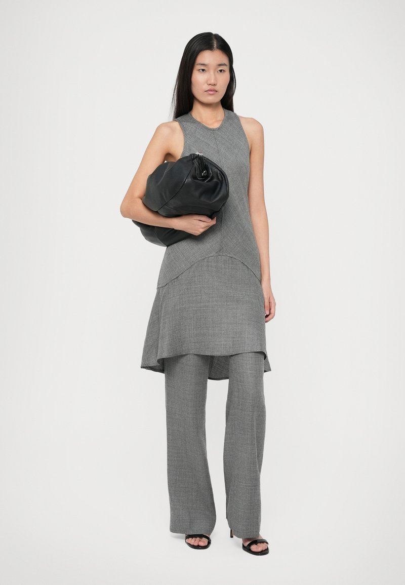 Femme portant un tunique sans manches grise avec un pantalon large assorti, tenant un grand sac à main noir et portant des talons ouverts noirs.