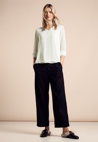 Blusa in menta chiaro con scollo a V e maniche tre quarti, abbinata a pantaloni neri larghi. Scarpe nere slip-on con dettagli in pelliccia.