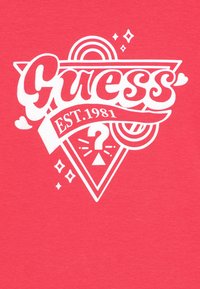 Červené bavlněné tričko s výrazným bílým nápisem "Guess", grafickým trojúhelníkem a stuhou s textem "EST.1981." Obsahuje detaily srdce a hvězdy.