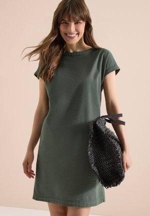Uśmiechnięta kobieta w zielonej, teksturowanej sukience z krótkim rękawem, trzymająca czarną plecioną torbę na jasnobeżowym tle.