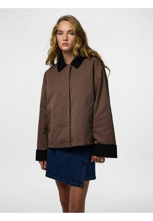 Ženska z dolgimi blond lasmi, oblečena v rjavo jakno s črnim ovratnikom in manšetami ter modro denim krilo, stoji pred enostavnim ozadjem.