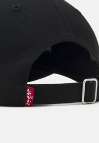 Casquette en coton noir avec un bord incurvé, dotée d'une étiquette de logo rouge et d'une boucle métallique ajustable à l'arrière pour le réglage de la taille.