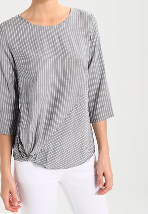 Femme portant un chemisier rayé verticalement gris et blanc avec des manches 3/4 et un détail noué à l'avant, associé à un pantalon blanc.