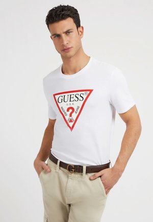 White cotton t-shirt featuring a red triangular logo with "GUESS" and additional text. Worn with beige trousers and a brown belt.