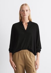 Maglietta nera a maniche lunghe con scollo a V e una piccola patta. Tessuto morbido e leggero con una vestibilità rilassata, abbinata a pantaloni beige.