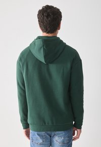Sudadera de algodón verde con capucha ajustable, puños y dobladillo acanalados; llevada sobre jeans azules, mostrando una textura suave y un diseño casual.