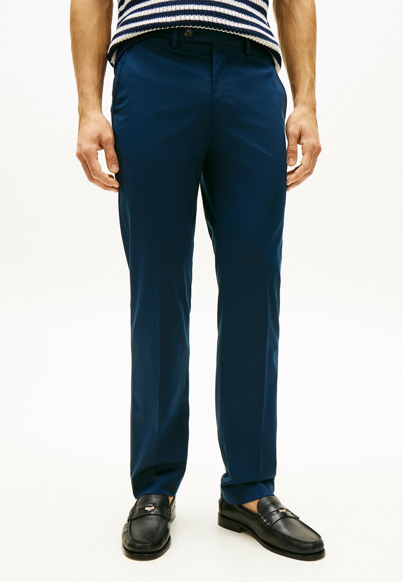 Homme portant un pantalon sur mesure bleu marine, des mocassins noirs et un pull en maille rayé bleu marine et blanc, debout devant un fond blanc.