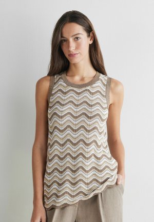Vrouw draagt een mouwloze beige en witte top met golvend patroon, staat met één hand in de zak tegen een effen lichte muur.