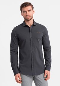 Donkergrijze langarmshirt met een knoopsluiting aan de voorkant, een scherpe kraag en een geborduurd logo op de borst. De stof lijkt glad en aansluitend.