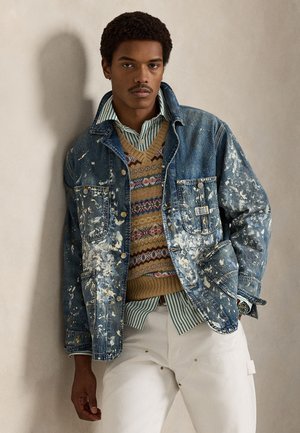 Mees, kes kannab värvipritsmetega teksajopet mustrilise kampsuni ja triibulise särgi kohal, valgete pükstega, seisab ühevärvilise seina ees.