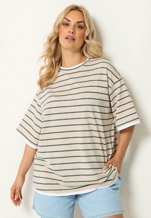 Blond kvinde med bølget hår bærer en beige og grønstribet oversize T-shirt og lyseblå denimshorts, stående mod en hvid baggrund.