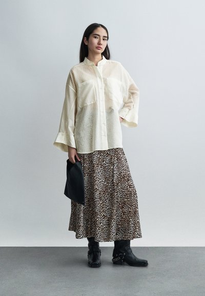 Διαφανές πουκάμισο σε εκρού με κουμπιά μπροστά και δύο μπροστινές τσέπες, συνδυασμένο με φούστα μάξι με λεοπάρ μοτίβο και μαύρα μποτάκια αστραγάλου. Κρατάει ένα μαύρο τσαντάκι χειρός.