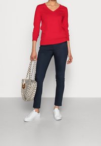 Pull rouge en V, pantalon slim-fit bleu marine, baskets blanches. Tenant un sac fourre-tout à motifs avec des couleurs claires et sombres. Textures lisses et ajustées.