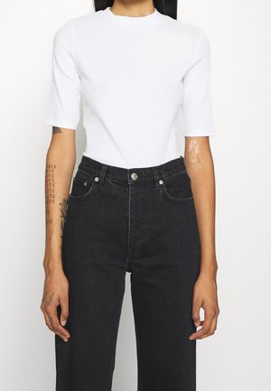 Persoon draagt een wit geribd topje met korte mouwen, ingestopt in een hoge taille zwarte spijkerbroek, staand tegen een effen achtergrond.