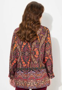 Long-sleeve patterned blouse featuring vibrant floral designs in reds, oranges, and purples, with a contrasting lower hem and button cuffs.