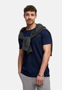 Navyblaues T-Shirt mit kurzen Ärmeln und einer Brusttasche, getragen mit einem grauen Sweatshirt über den Schultern und grauen Hosen.