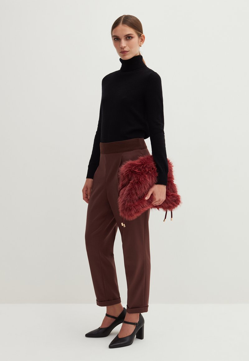 Schwarzer Rollkragenpullover, braune hochgeschnittene Hose mit umgeschlagenen Säumen und schwarze Pumps. Hält eine rote Tasche aus Kunstfell mit goldenen Akzenten.
