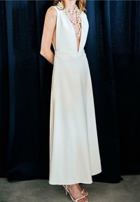 Femme portant une robe blanche sans manches avec un col en V profond, des colliers de perles superposés et des talons blancs ouverts, debout devant un rideau sombre.