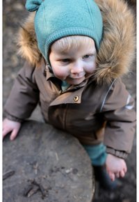 Brown outer jacket with faux fur hood, green knit hat, and blue undergarment; child rests hand on a wooden log surface outdoors.
