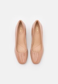 Escarpins en cuir verni nude avec un bout arrondi, une texture lisse et une semelle intérieure rembourrée. Logo doré "Clarks" visible sur la doublure intérieure.