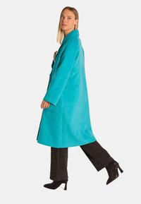 Türkisfarbener Wollmischungs-Mantel mit strukturiertem Design, einreihigem Verschluss und Reverskragen; kombiniert mit schwarzen, genagelten Hosen und Absatzstiefeln.