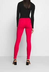 Jeans ajustados en denim rosa vibrante. Presentan una cintura alta, dos bolsillos traseros y detalles de costura sutiles. Combinados con sandalias negras de tacón alto.