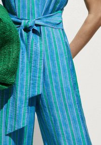 Salopette a righe blu e verdi con cintura annodata in vita, tessuto strutturato, abbinata a una borsa intrecciata verde.